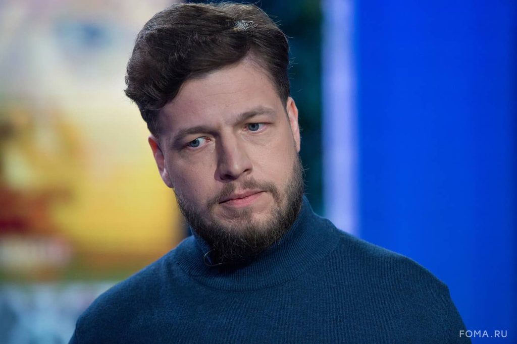 поэт Алексей Шмелев. Он запечатлён в полуанфас: взгляд чуть отведён в сторону, выражение лица спокойное, с едва заметной полуулыбкой. У Алексея аккуратная тёмно‑каштановая причёска с лёгкой волной, ухоженная борода и усы. Он одет в тёмно‑синий свитер с высоким воротом — лаконичный образ подчёркивает сдержанную эстетику кадра. Фон размытый: слева — тёплые жёлто‑оранжевые пятна, справа — насыщенный синий цвет с вертикальной светлой линией; контраст создаёт студийную атмосферу