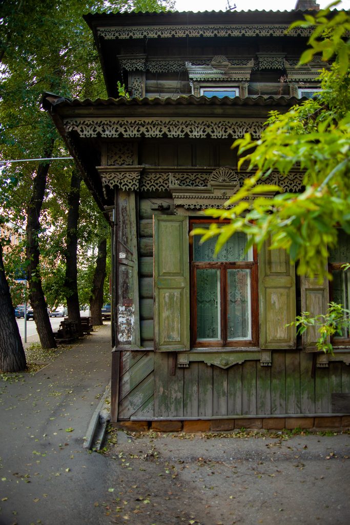 На фото — фрагмент старинного деревянного дома, характерного для исторической застройки Иркутска. Здание выполнено в традициях сибирского деревянного зодчества: сруб из брёвен дополнен выразительным резным декором.
В центре кадра — окно с тёмно‑коричневой деревянной рамой и узорчатыми стёклами; по бокам — выцветшие зелёные ставни с филёнками, местами облупившаяся краска подчёркивает возраст постройки. Над окном — сложный резной карниз с ажурным орнаментом: пропильная резьба образует «кружевной» узор, а накладные элементы добавляют объём. В декоре угадываются растительные и геометрические мотивы — типичный приём иркутских мастеров конца XIX — начала XX века.
Нижняя часть фасада обшита досками с фигурной отделкой; на древесине заметны следы времени — потемнения, налёт зеленоватой патины, потёртости. Свес крыши оформлен волнистым металлическим профилем, который контрастирует с тонкой работой резчиков.