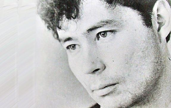 На фото — чёрно‑белый портрет Александра Вампилова, выполненный в стилистике документальной съёмки середины XX века. В кадре — крупный план лица драматурга: он слегка повернул голову вправо, взгляд устремлён чуть выше уровня объектива — в нём читается сосредоточенность и лёгкая задумчивость. У Вампилова выразительные черты: прямой нос, чётко очерченные скулы, плотно сомкнутые губы. Тёмные брови подчёркивают глубину взгляда. Причёска — волнистые волосы, уложенные небрежно, с завитками у лба; пряди мягко обрамляют лоб и виски. Текстура кожи передана с детализацией: заметны едва уловимые тени, подчёркивающие рельеф лица. Фон нейтральный, светлый, без узоров — лёгкая зернистость снимка придаёт ему винтажный характер. Освещение мягкое, рассеянное: оно формирует плавные переходы света и тени, не создавая резких контрастов.