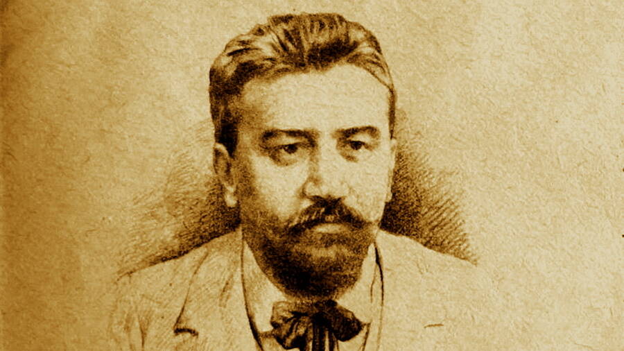 На фото — портрет Александра Ивановича Куприна, выполненный в стилистике старой фотографии или гравюры: тёплый сепиевый тон придаёт изображению историческую глубину.
Писатель изображён по грудь, взгляд направлен чуть в сторону от камеры — в нём читается спокойная сосредоточенность. У Куприна аккуратная причёска с пробором, густые тёмные волосы гладко зачёсаны назад. Выразительные черты лица подчёркивают пышные усы и плотная борода.
На Александре Ивановиче — светлый костюм, под ним видна рубашка с жёстким воротником и тёмный галстук‑бабочка. Фон нейтральный, без деталей: лёгкая штриховка создаёт эффект студийного задника, акцентируя всё внимание на образе писателя.